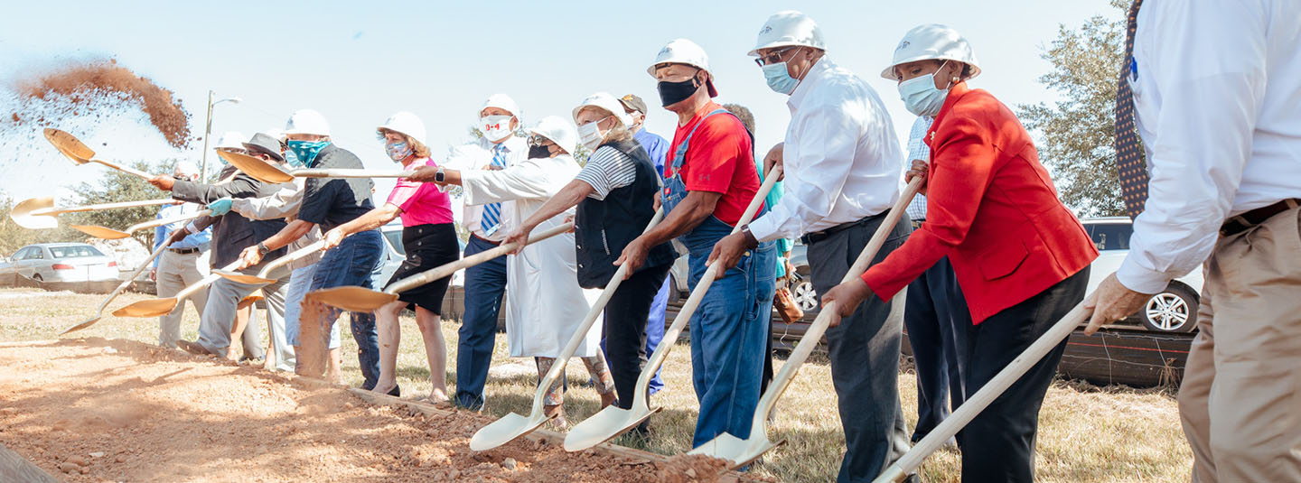 A group of thirteen men and women in a line with shovels and wearing hardhats at a ground breaking for a new construction project