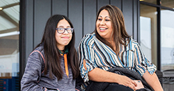A teen with glasses smiling sitting beside her mother in front of a school building